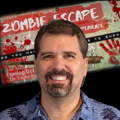 Smiling man with a goatee in front of a Zombie Apocalypse poster